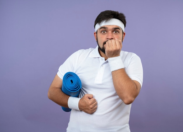 Betroffener junger sportlicher Mann, der Stirnband und Armband hält, die Yogamatte hält, die Hand auf Mund lokalisiert auf grüner Wand mit Kopienraum setzt