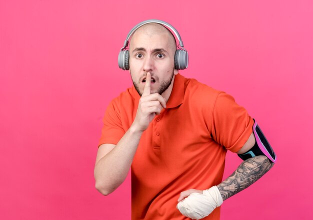Betroffener junger sportlicher Mann mit Handgelenksverband, der Kopfhörer mit Telefonarmband trägt, das Stille Geste zeigt, die auf rosa Wand isoliert wird