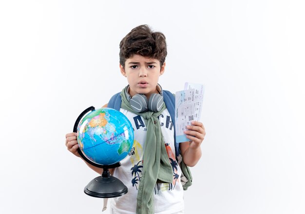 Betroffener kleiner Schuljunge, der Rückentasche und Kopfhörer trägt, die Tickets und Globus lokalisiert auf weißem Hintergrund halten