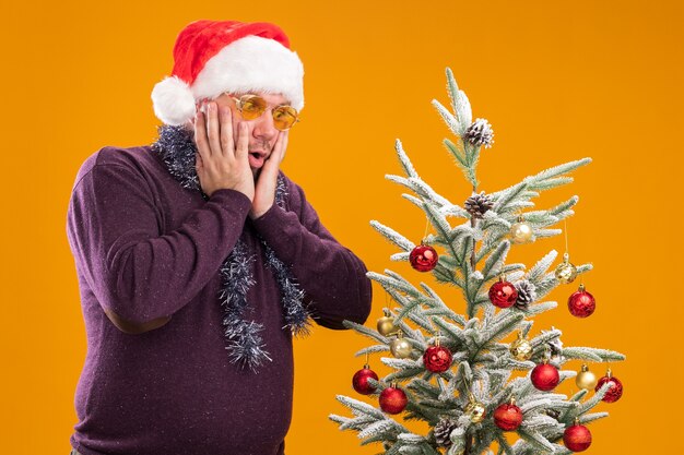 Betroffener Mann mittleren Alters mit Weihnachtsmütze und Lametta-Girlande um den Hals mit Brille, die in der Nähe des geschmückten Weihnachtsbaums steht und die Hände im Gesicht hält