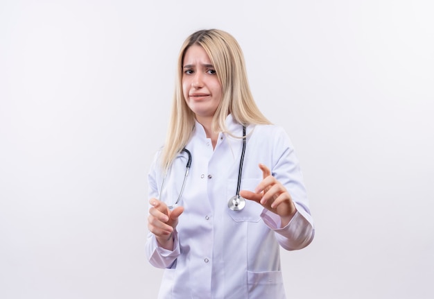 Betroffenes junges blondes Mädchen des Arztes, das Stethoskop und medizinisches Kleid trägt, das Größe mit beiden Händen auf isolierter weißer Wand zeigt