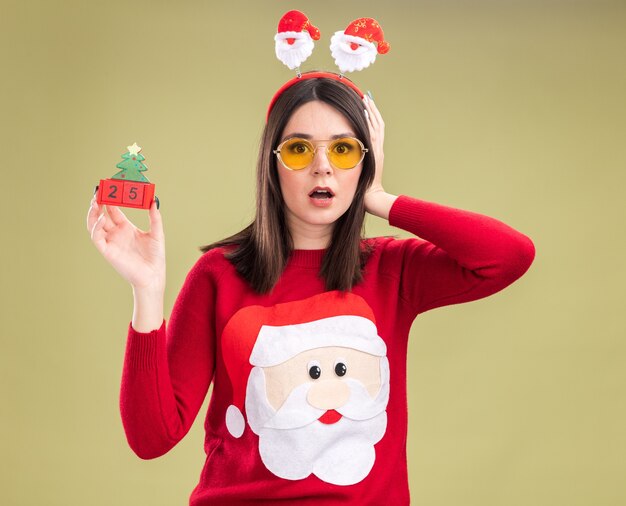 Betroffenes junges hübsches kaukasisches Mädchen, das Weihnachtsmann-Pullover und Stirnband mit Brille trägt, die Weihnachtsbaumspielzeug mit Datum halten