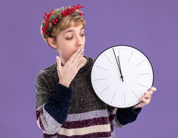 Betroffenes junges hübsches Mädchen, das Weihnachtskopfkranz hält und Uhr hält, die Hand auf Mund lokalisiert auf lila Hintergrund hält