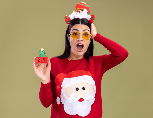 Betroffenes junges kaukasisches Mädchen, das Weihnachtsmann-Stirnband und Pullover mit Gläsern hält, die Weihnachtsbaumspielzeug mit Datum halten Hand auf Kopf lokalisiert auf olivgrüner Wand halten