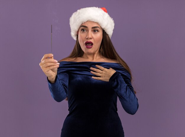 Betroffenes junges schönes Mädchen, das blaues Kleid und Weihnachtsmütze hält und Wunderkerzen hält und Hand sich lokalisiert auf lila Wand setzt