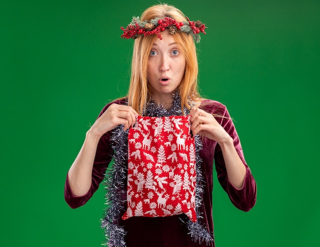 Kostenloses Foto betroffenes junges schönes mädchen, das rotes kleid mit kranz und girlande am hals hält, die weihnachtstasche lokalisiert auf grüner wand trägt