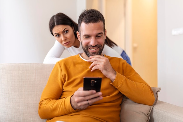 Betrüger Mann, der online mit einem Smartphone und einer Freundin datiert, spioniert das Sitzen auf einem Sofa zu Hause aus
