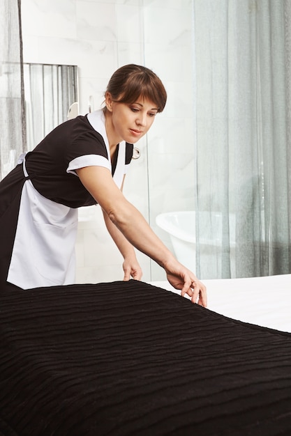 Kostenloses Foto bett machen ist wie kunst. innenaufnahme eines dienstmädchens in uniform, das beim reinigen der hotelwohnung oder des hauses der eigentümer eine decke auf das bett legt und versucht, staub von allen oberflächen abzuwischen und besten service zu bieten