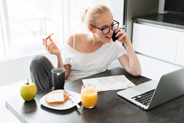 Beuatiful Dame, die mit Laptop arbeitet und auf Smartphone spricht und Frühstück isst