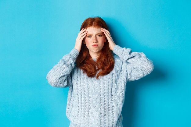 Beunruhigtes rothaariges Mädchen im Teenageralter, das den Kopf berührt, mit unruhigem Gesichtsausdruck schaut und vor blauem Hintergrund steht, hat ein Problem.