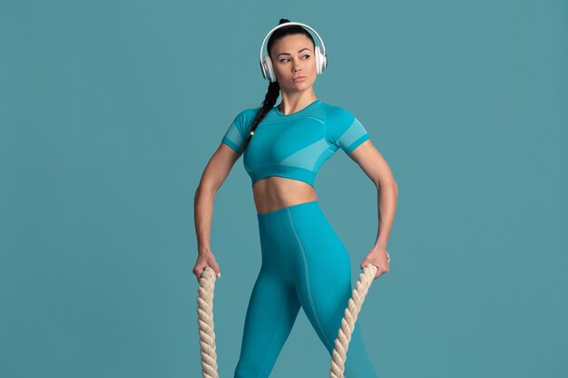 Bewegung. Schöne junge Sportlerin üben, einfarbiges blaues Porträt. Brünettes Modell mit sportlicher Passform mit Seilen. Bodybuilding, gesunder Lebensstil, Schönheits- und Aktionskonzept.