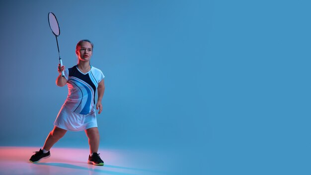 Bewegung. Schöne Zwergfrau, die im Badminton übt, isoliert auf Blau im Neonlicht
