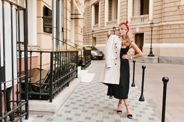 Bezaubernde blonde Frau in stilvollem schwarzen Kleid und Schuhen mit hohen Absätzen, die morgens im Freien posieren. Schönes blondes Mädchen in eleganter Kleidung, das Zeit auf der Straße verbringt und lächelt.