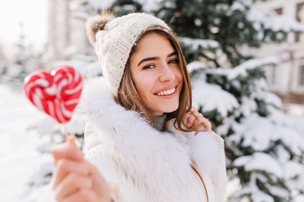 Bezaubernde freudige junge Frau des Winternahaufnahmeporträts im sonnigen Wintermorgen mit rosa lollypop auf Straße