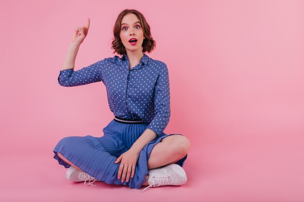 Bezaubernde junge Dame in blauer Kleidung, die mit überraschtem Gesichtsausdruck auf dem Boden sitzt. Innenporträt des brünetten charmanten Mädchens im Rock.