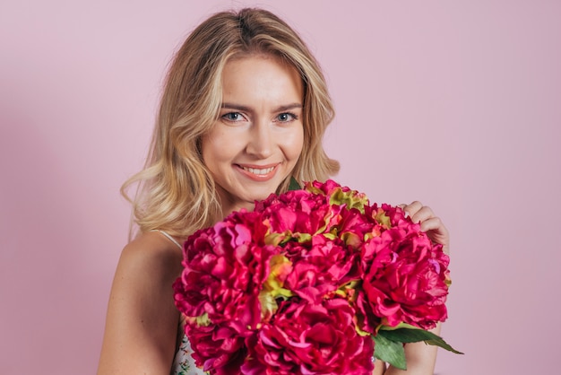 Bezaubernde lächelnde blonde junge Frau, die Blumenblumenstrauß gegen rosa Hintergrund hält
