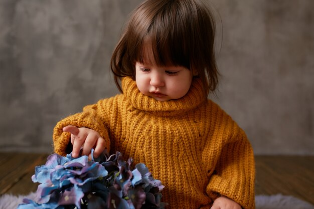 Bezauberndes kleines Mädchen in der orange Strickjacke erforscht die blauen Hortensien, die auf warmer Decke sitzen