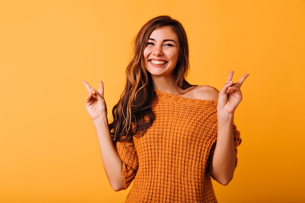 Bezauberndes Mädchen mit langer Frisur, die mit Friedenszeichen auf Orange aufwirft. Frohes weibliches Modell im trendigen Strickpullover, der im Studio lacht.