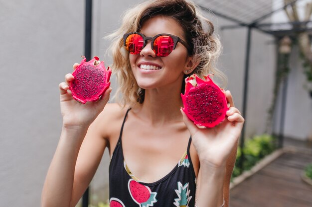 Bezauberndes Mädchen trägt rosa Sonnenbrille mit saftiger Pitaya. Porträt der charmanten lächelnden Frau, die exotische Früchte isst.