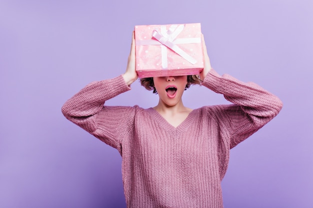 Bezauberndes mädchen trägt wollpullover isoliert auf lila wand mit rosa geschenkbox. innenfoto der glücklichen kurzhaarigen frau, die spaß mit neujahrsgeschenk hat.