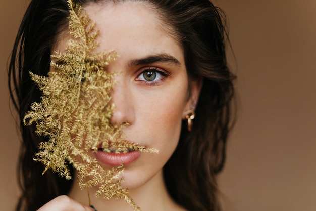 Bezauberndes weißes Mädchen mit trendigem Schmuck, der mit Pflanze aufwirft. Nahaufnahme des verblüfften weiblichen Modells mit goldenem Zubehör und grünem Blatt.