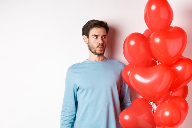 Beziehung. Junger Mann, der verwirrt auf Herzballon schaut, verwirrt am Valentinstag, stehend auf weißem Hintergrund
