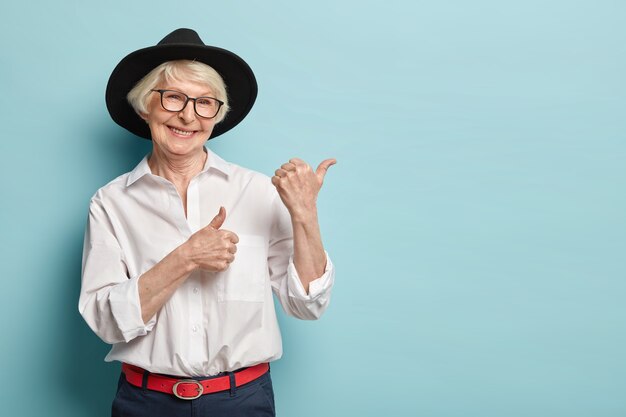 Bild der attraktiven faltigen Frau mit ansprechendem Blick, fühlt sich erfrischt, jung für ihr Alter, zeigt in der oberen rechten Ecke, zufrieden mit dem Produkt, trägt weißes Hemd, schwarze Kopfbedeckung, optische Brille