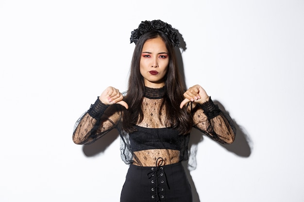 Bild der enttäuschten asiatischen Frau im Halloween-Kleid des gotischen untoten Mädchens, das Daumen nach unten zeigt, etwas Schlechtes nicht mag und nicht damit einverstanden ist und über weißem Hintergrund steht.