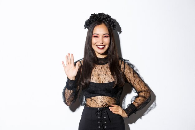 Bild der freundlichen stilvollen asiatischen Frau im gotischen Spitzenkleid, das Hand winkt, um Hallo zu sagen, jemanden zu begrüßen, Leute auf Halloween-Party willkommen zu heißen, die über weißem Hintergrund stehen.