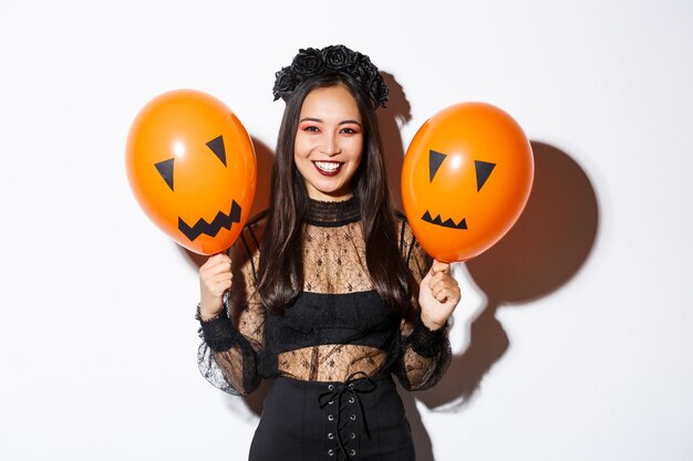 Bild der fröhlichen asiatischen Frau im Hexenkostüm, das Halloween feiert und Luftballons mit unheimlichen Gesichtern hält, die über weißem Hintergrund stehen.