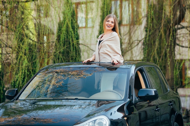 Bild der fröhlichen jungen Frau, die Sonnenbrille und erhobene Hände auf dem Schiebedach des Luxusautos trägt