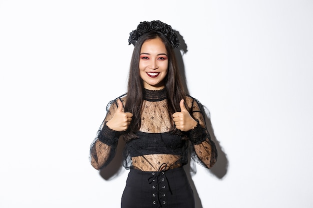 Bild der glücklichen asiatischen Frau im Halloween-Partykleid, das Daumen hoch in Zustimmung zeigt. Mädchen, das als Hexe oder Witwe trägt, die erfreut sieht, wie oder stimmt mit etwas, weißem Hintergrund überein.