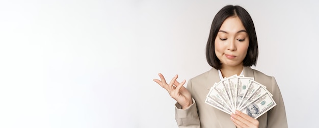 Bild der glücklichen Geschäftsfrau der asiatischen Unternehmensfrau, die Geldbargelddollar zeigt und das Denken im Anzug über weißem Hintergrund steht