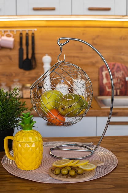 Kostenloses Foto bild der großen hellen küche mit weißen und braunen schränken mit gelbem ananas-teekessel, weißer pfeffermühle und metall, das mit früchten hängt