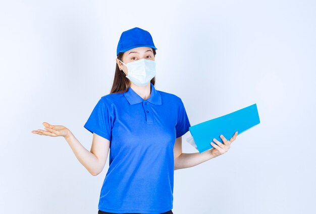 Bild der jungen Frau in medizinischer Maske, die mit blauem Ordner über weißer Wand posiert.