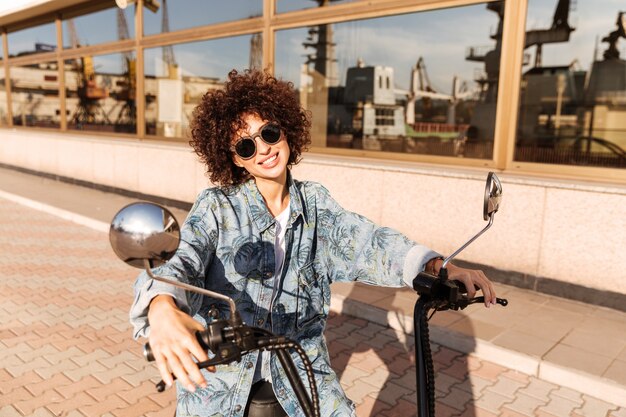 Bild der lächelnden lockigen Frau in der Sonnenbrille, die auf Motorrad sitzt