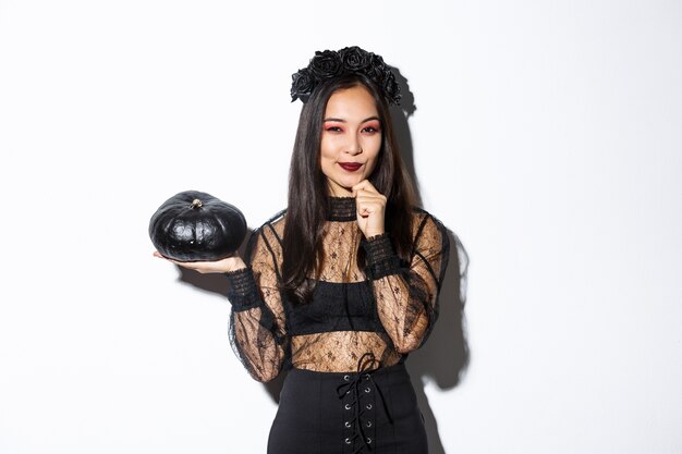 Bild der nachdenklichen lächelnden asiatischen Hexe, Mädchen im Halloween-Kostüm, das Kamera betrachtet und schwarzen Kürbis zeigt.