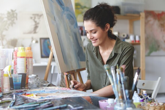 Bild der niedlichen Künstlerin, die am Tisch sitzt, umgeben von Aquarellen, etwas an der Staffelei zeichnend, glücklichen Ausdruck habend. Brünette junge Frau, die mit kreativer Arbeit an Werkstatt beschäftigt ist