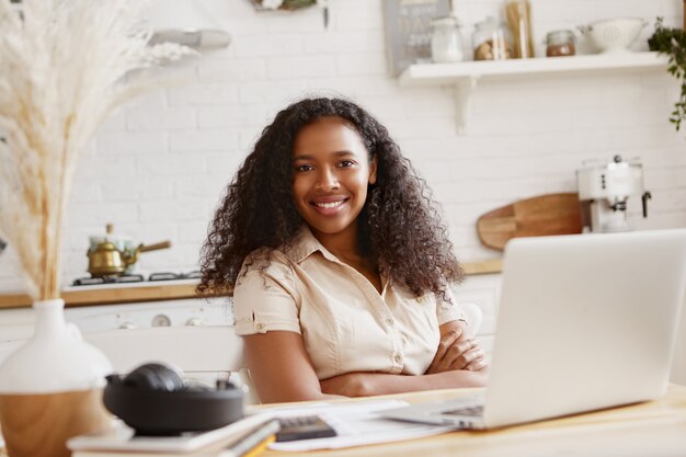 Bild der niedlichen stilvollen jungen afroamerikanischen Buchhalterin mit zuversichtlichem zahnigem Lächeln, das entfernt auf Laptop-Computer arbeitet und Finanzen in der Küche tut. Technologie, Beruf und freiberuflich tätig