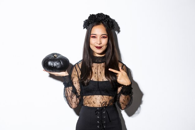 Bild der schönen asiatischen Frau im gotischen Spitzenkleid und im Kranz, die Finger auf schwarzen Kürbis zeigen, Halloween feiern, Hexenkostüm tragend, über weißem Hintergrund stehend.