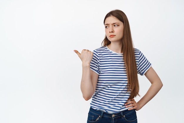 Bild der skeptischen jungen Frau, die mit zweifelhaftem, unbeeindrucktem Gesichtsausdruck die Stirn runzelt, zeigt und nach links schaut und auf Weiß steht.