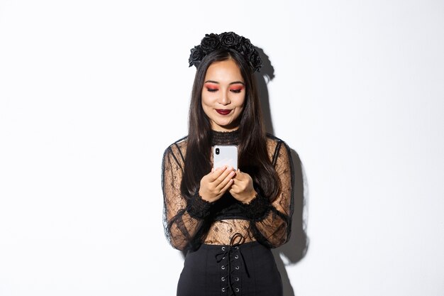 Bild der stilvollen asiatischen Frau im Halloween-Kostüm, das Nachrichten auf Handy überprüft. Mädchen im gotischen Spitzenkleid, das Smartphone betrachtet, über weißem Hintergrund stehend.