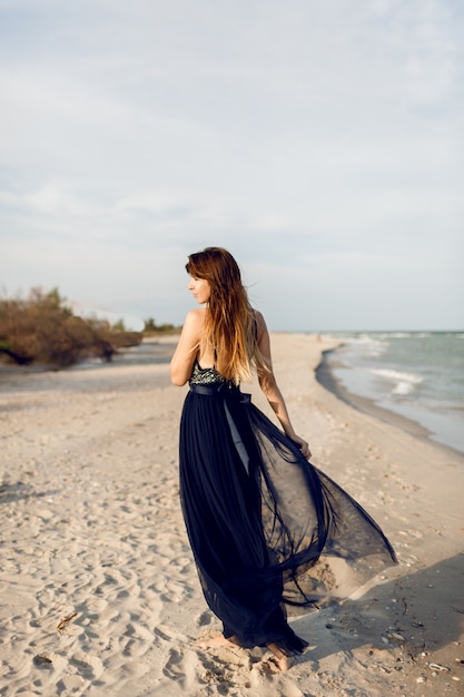 Bild der vollen Höhe der modischen Frau im eleganten Luxuskleid, das am Strand aufwirft. Blick von hinten. Lange Haare.