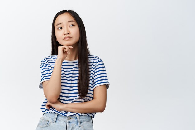 Bild des asiatischen Mädchens denkt, schaut mit nachdenklichem Gesichtsausdruck beiseite, entscheidet etw, steht auf Weiß. Platz kopieren