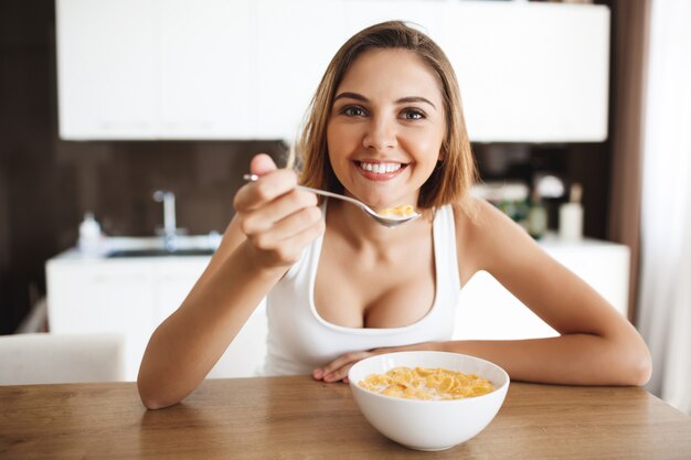 Bild des attraktiven jungen Mädchens, das Cornflakes mit Milch am lächelnden Küche isst