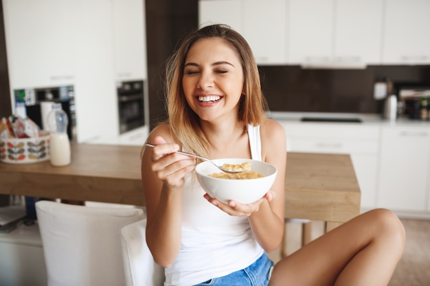 Bild des attraktiven jungen Mädchens, das lacht, während Cornflakes mit Milch an der Küche essen