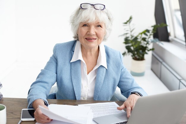 Bild des attraktiven selbstbewussten älteren reifen weiblichen Finanzberaters mit dem kurzen grauen Haar, das mit dem Lächeln schaut und ein Stück Papier in ihren Händen studiert, während sie an ihrem Schreibtisch arbeitet