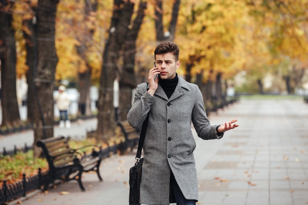 Bild des eleganten Mannes im Mantel mit Tasche gehend in Stadtpark und auf Smartphone im Herbst sprechend