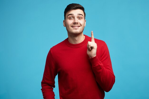 Bild des emotionalen unrasierten jungen europäischen Mannes im roten Pullover, der Zeigefinger hebt, um Ihre Aufmerksamkeit zu ziehen