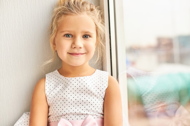 Bild des entzückenden hübschen Vorschulmädchens mit den großen blauen Augen, die schönes Kleid mit aufgeregtem glücklichem Lächeln tragen, Freunde auf ihrer Geburtstagsfeier suchend, am Fenster sitzend